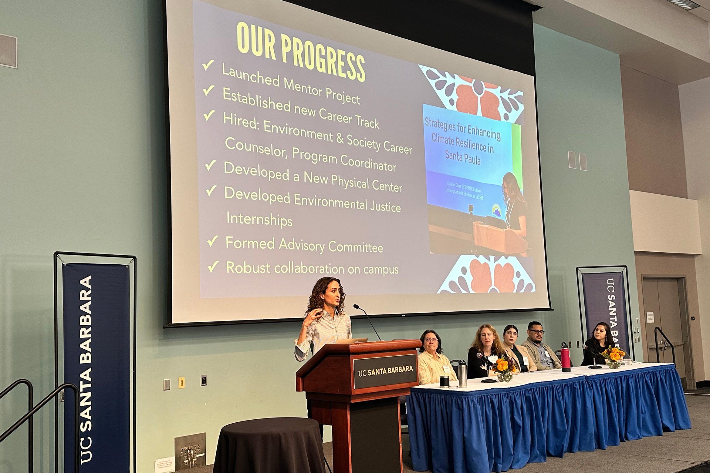 person standing at a podium in front of a projector screen and next to a panel of other seated people